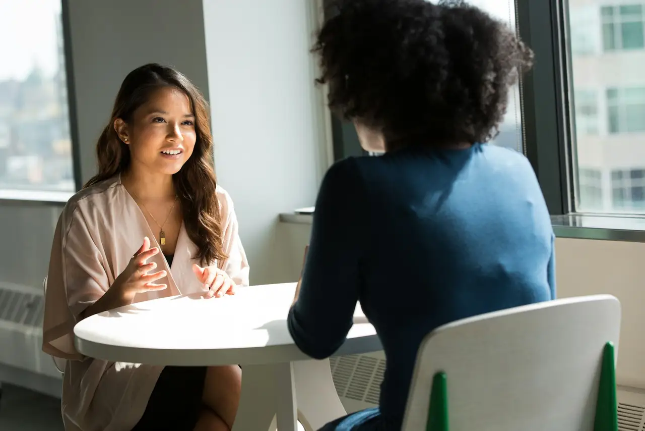 Two colleagues in conversation across a small table in a bright office.